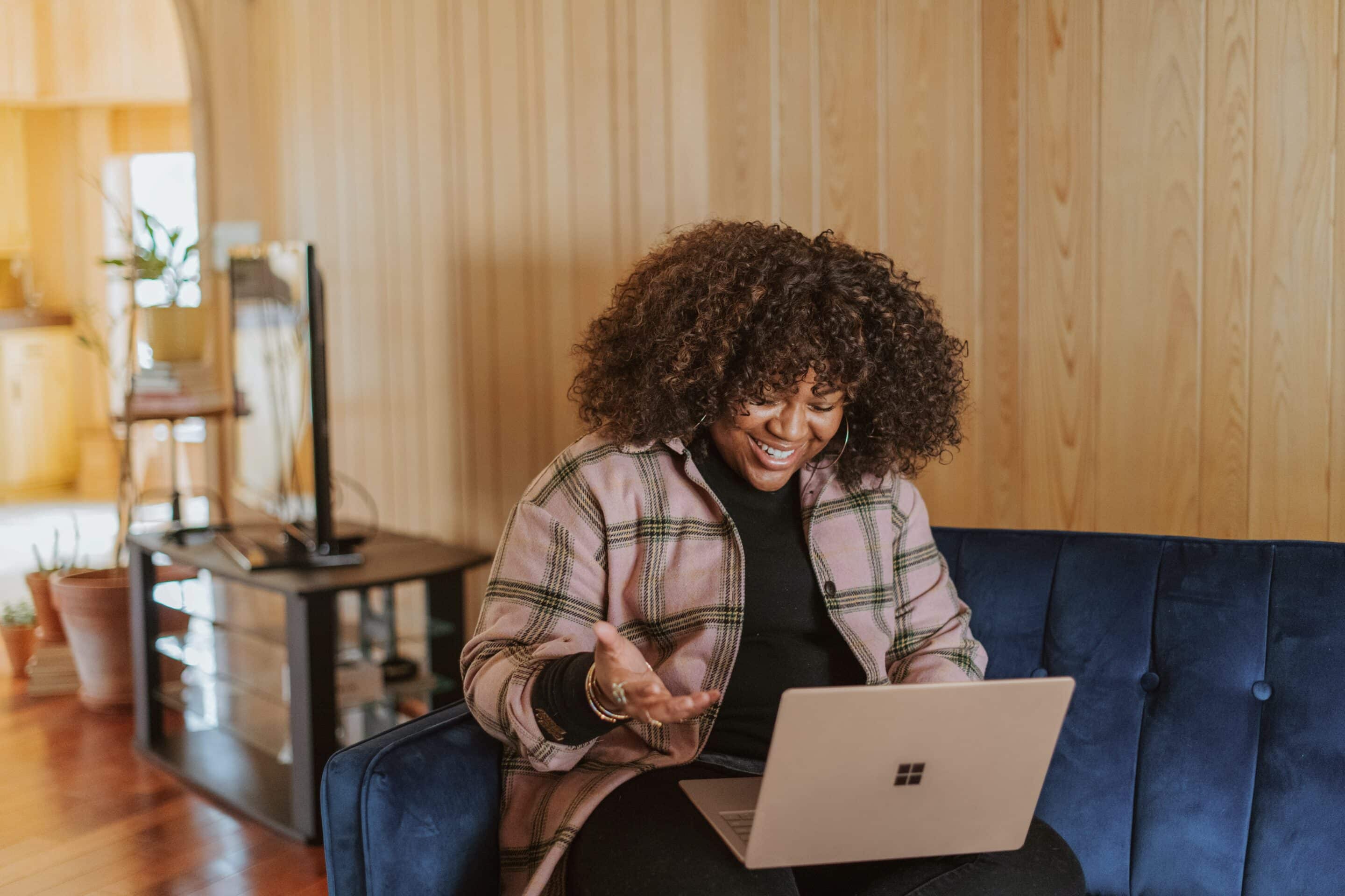 A black woman with curly hair sitting on a blue sofa, smiling at her laptop.